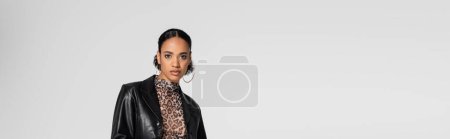 stylish african american woman in trendy leather jacket and hoop earrings isolated on grey, banner 