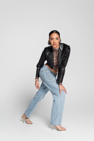 full length of fashionable african american woman in high heeled sandals and trendy cropped jacket posing on grey 