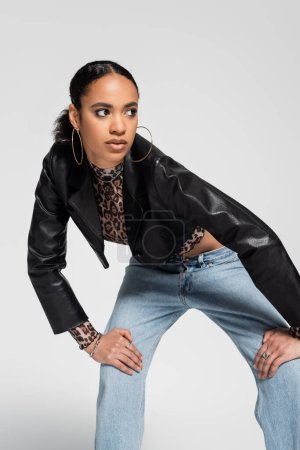 fashionable african american woman in trendy cropped jacket and blue jeans looking away while posing isolated on grey 