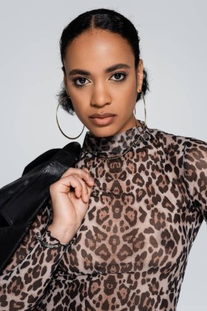portrait of fashionable african american woman in cropped top with animal print holding black jacket isolated on grey 