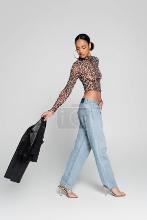 Photo for Full length of well dressed african american model in crop top with animal print holding black jacket and walking on grey - Royalty Free Image