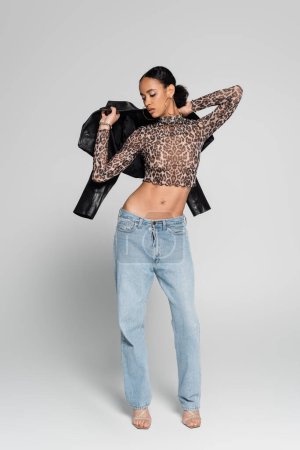 full length of young african american model in crop top with animal print holding black jacket on grey 
