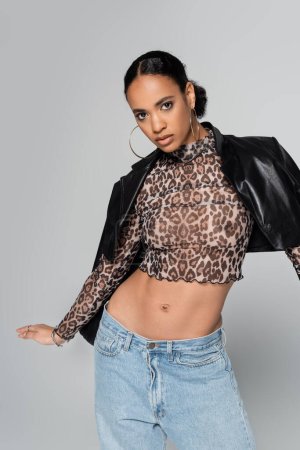 fashionable african american woman in crop top with animal print and leather jacket on shoulders posing isolated on grey 