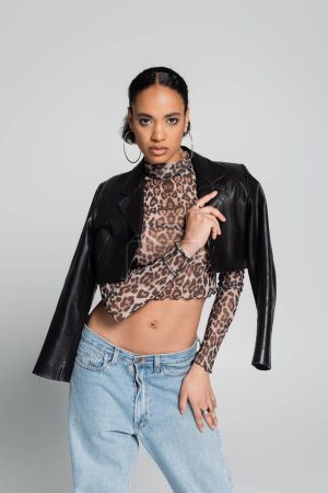 stylish african american model in crop top with animal print wearing black leather jacket isolated on grey 