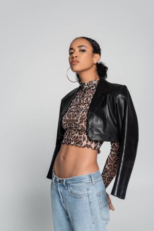 stylish african american model in crop top with animal print and black leather jacket posing isolated on grey 