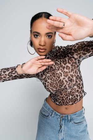 stylish african american woman in crop top with animal print and denim jeans gesturing isolated on grey 