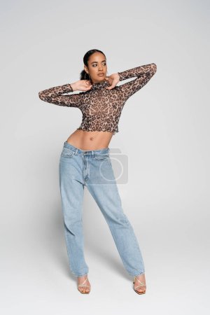 full length of brunette african american woman in crop top with animal print and denim jeans posing on grey 