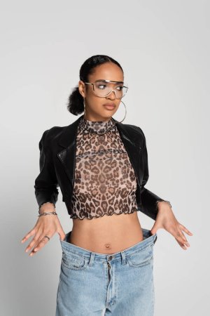 slim african american woman in cropped jacket and trendy sunglasses posing isolated on grey 