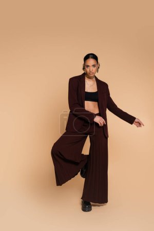 full length of young african american woman in stylish suit with wide pants standing on one leg while posing on beige 
