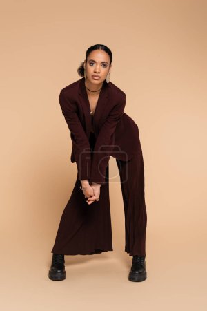 full length of young african american woman in maroon blazer and wide pants posing on beige 