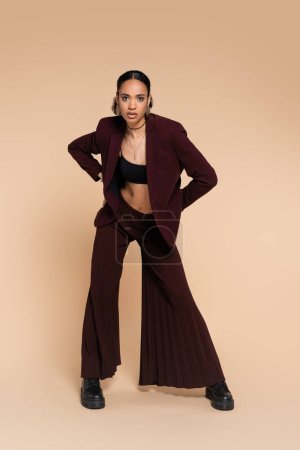 full length of young african american woman in stylish blazer and maroon wide pants posing with hands on hips on beige 