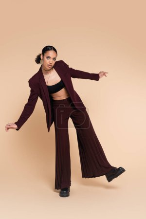 full length of pretty african american woman in maroon suit with wide leg pants posing on beige 