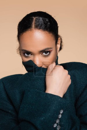 portrait of young african american woman covering face with collar of oversized blazer isolated on beige 