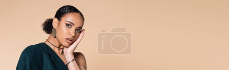 Téléchargez les photos : Jeune modèle afro-américain en blazer bleu foncé touchant le visage tout en posant isolé sur beige, bannière - en image libre de droit
