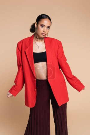 stylisches afrikanisch-amerikanisches Model in Crop Top und rotem Blazer posiert isoliert auf beige 
