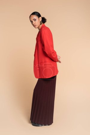 full length of stylish african american model in wide leg pants and red blazer posing on beige 