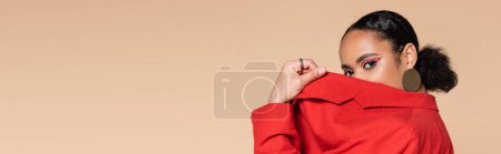 Téléchargez les photos : Portrait de brune modèle afro-américain en blazer rouge vif couvrant le visage tout en posant isolé sur beige, bannière - en image libre de droit