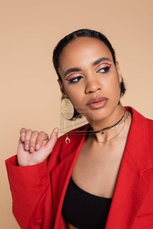 Foto de Retrato de modelo afroamericano con estilo en blazer rojo brillante tirando de collar de cadena de oro aislado en beige - Imagen libre de derechos