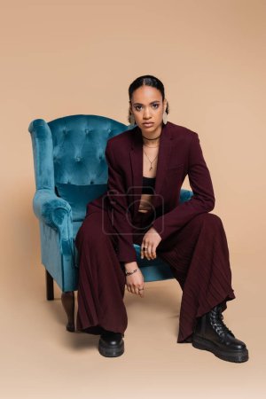 Photo for Stylish african american model in maroon suit sitting on blue velvet armchair on beige - Royalty Free Image