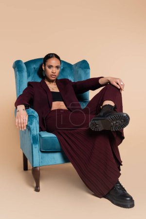 stylish african american model in maroon blazer and wide pants sitting on blue velvet armchair on beige 