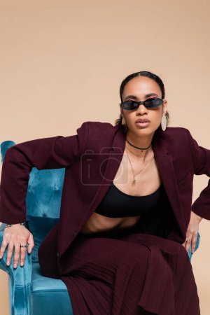 african american model in stylish suit and trendy sunglasses posing on blue velvet armchair on beige 