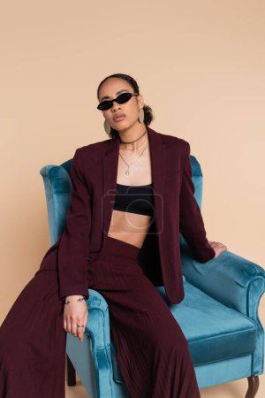 Photo for African american model in burgundy suit and stylish sunglasses sitting on blue velvet armchair on beige - Royalty Free Image