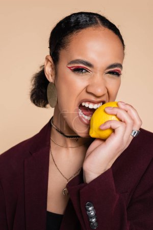 Afroamerikanerin im weinroten Blazer beißt in reife Zitrone auf Beige 