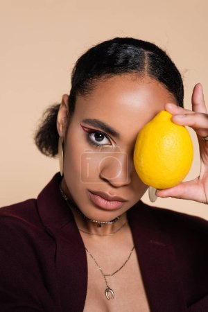 Photo for African american woman in burgundy blazer holding ripe lemon near eye isolated on beige - Royalty Free Image