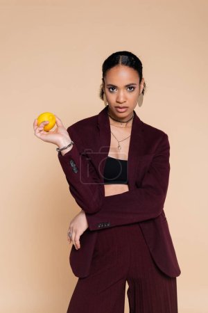 Téléchargez les photos : Femme afro-américaine en blazer bordeaux posant avec du citron frais isolé sur beige - en image libre de droit