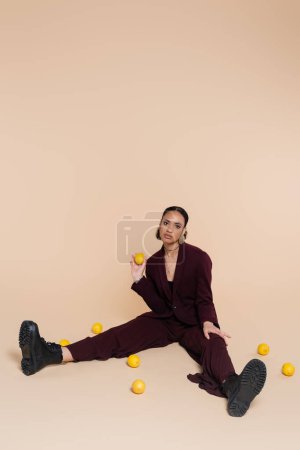 Téléchargez les photos : Élégante femme afro-américaine en costume bordeaux assis autour de citrons frais sur beige - en image libre de droit
