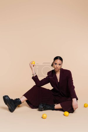 Foto de Modelo afroamericano con estilo en traje marrón sentado alrededor de limones frescos en beige - Imagen libre de derechos