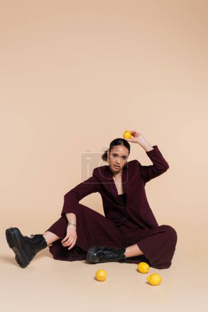 young african american woman in stylish burgundy suit sitting around fresh lemons on beige 
