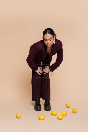 stylische afrikanisch-amerikanische Frau im weinroten Anzug steht und auf frische Zitronen auf Beige blickt 
