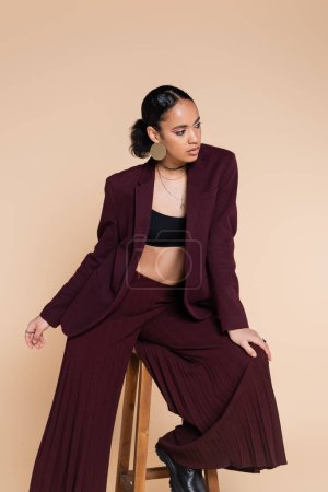 brunette african american woman in stylish burgundy suit sitting on wooden high stool while posing isolated on beige 