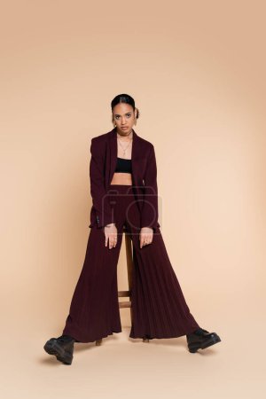 full length of stylish african american woman in burgundy suit with wide pants standing near wooden high chair on beige 