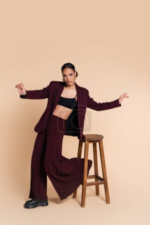 full length of stylish african american woman in burgundy suit with wide pants posing near wooden high stool on beige 