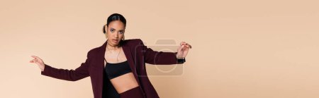 stylish african american woman in burgundy blazer and crop top posing isolated on beige, banner 