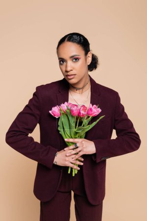 fashionable african american woman in burgundy suit posing with pink tulips isolated on beige 
