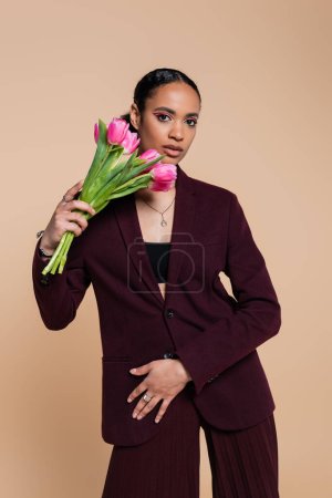 well dressed african american woman in burgundy suit posing with tulips isolated on beige 