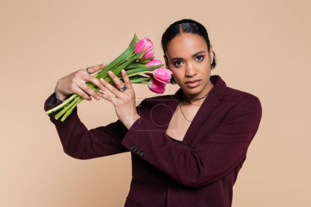 Photo for Well dressed african american woman in maroon blazer posing with pink tulips isolated on beige - Royalty Free Image