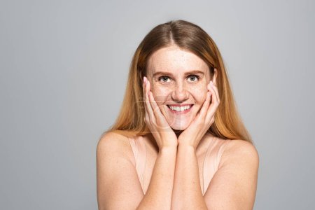 Cheerful freckled woman looking at camera and touching cheeks isolated on grey 