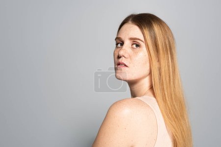 Young woman with freckled skin looking at camera isolated on grey 