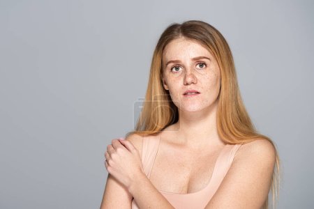 Young model with freckles on face touching shoulder isolated on grey 