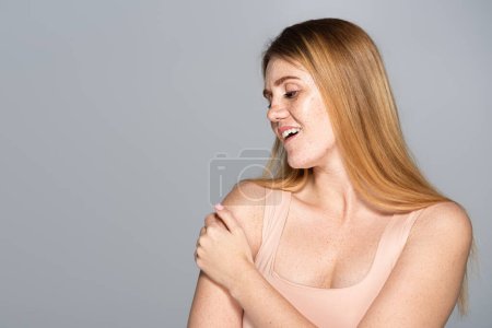 Smiling woman with freckled skin looking at shoulder isolated on grey 