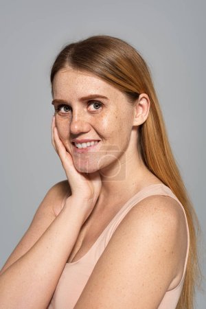 Smiling woman with freckles on face and body touching cheek isolated on grey 