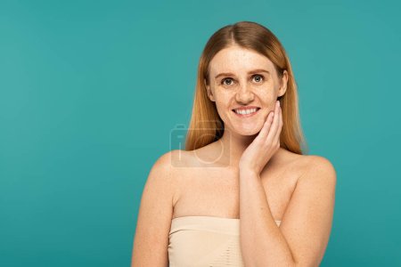 Smiling woman with freckles and naked shoulders posing isolated on turquoise