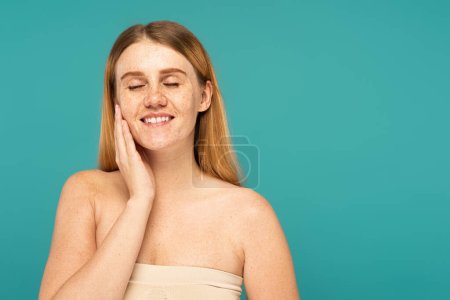 Smiling woman closing eyes while touching freckled skin isolated on turquoise
