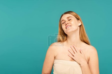 Positive woman closing eyes and touching freckled skin isolated on turquoise
