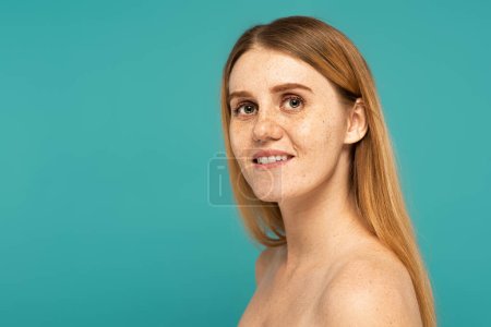Positive woman with freckled skin and naked shoulders standing isolated on turquoise