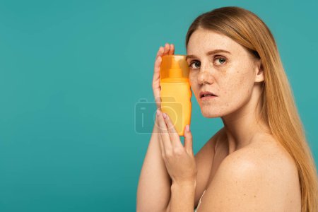 Freckled woman holding sunscreen and looking at camera isolated on turquoise